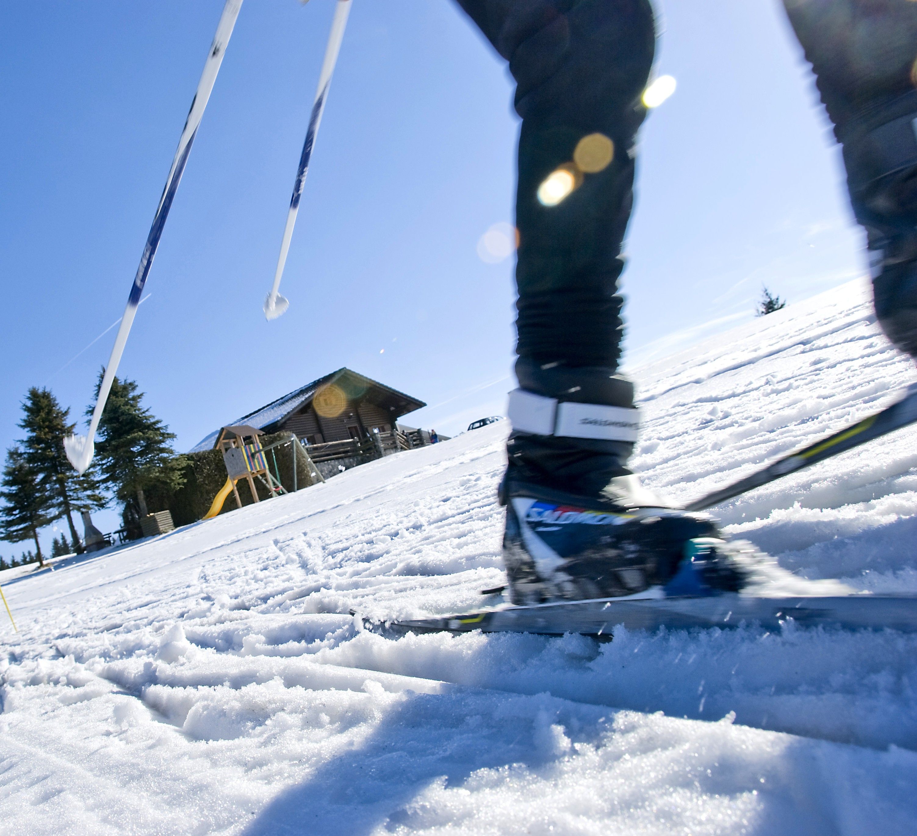 Nahaufnahme eines Langläufers auf einer schneebedeckten Piste mit einer Holzhütte im Hintergrund.