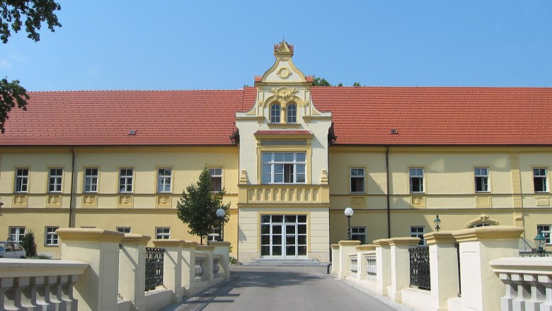 Gelbes Gebäude mit rotem Dach und dekorativer Fassade, umgeben von einer Brücke und Bäumen.