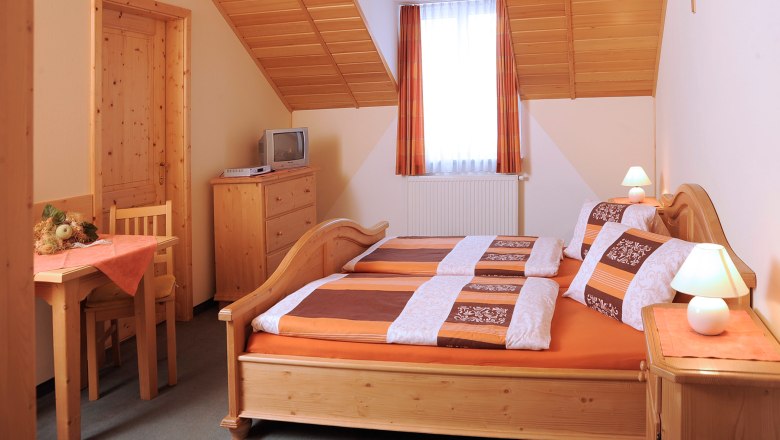 Gem&uuml;tliches G&auml;stezimmer mit Holzm&ouml;beln, Doppelbett, kleinem Tisch, Fernseher und Dachschr&auml;ge.