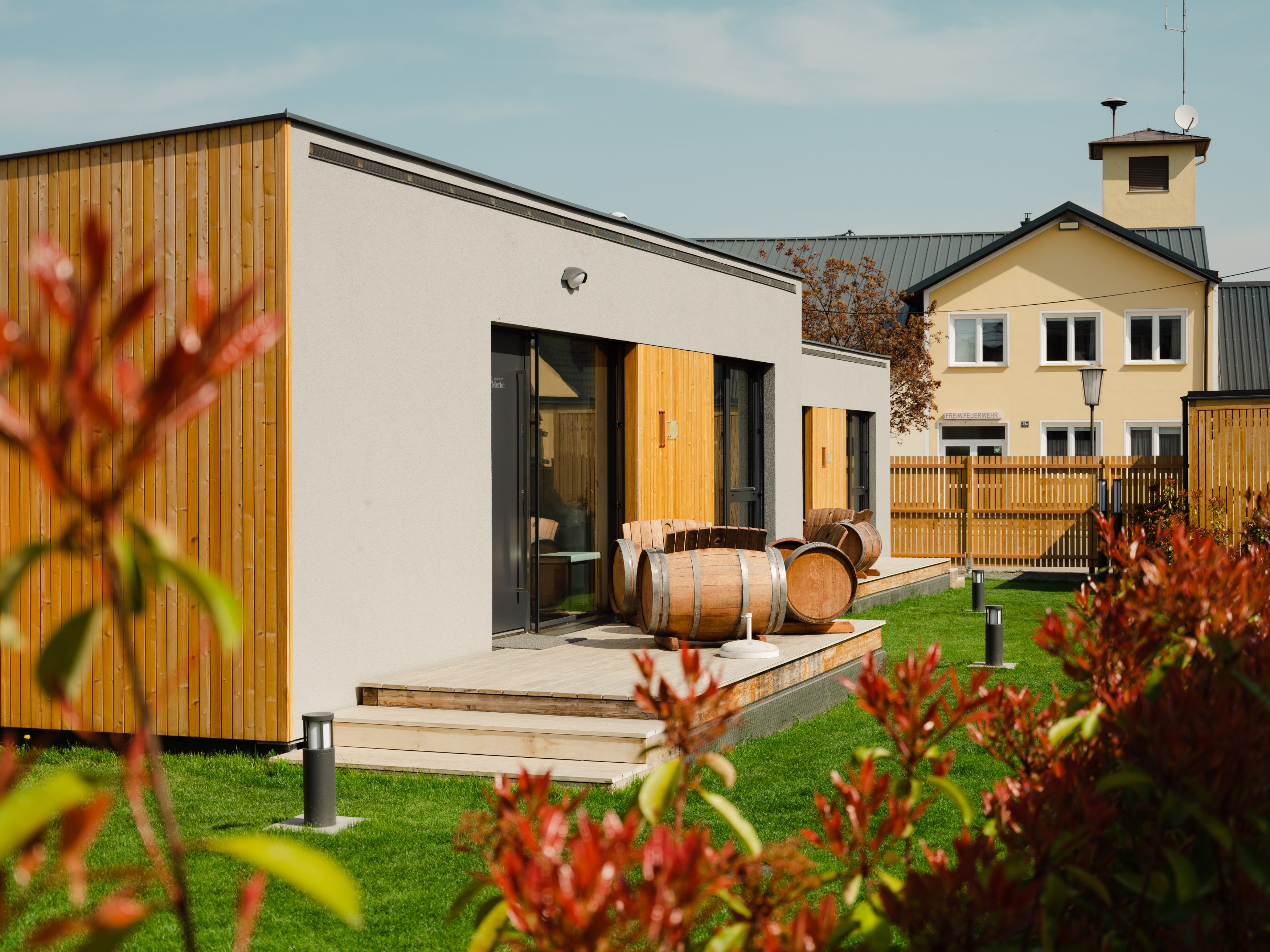 Moderne Appartements mit Holzverkleidung und Weinfässern auf der Terrasse, umgeben von grünem Rasen und einem Zaun.