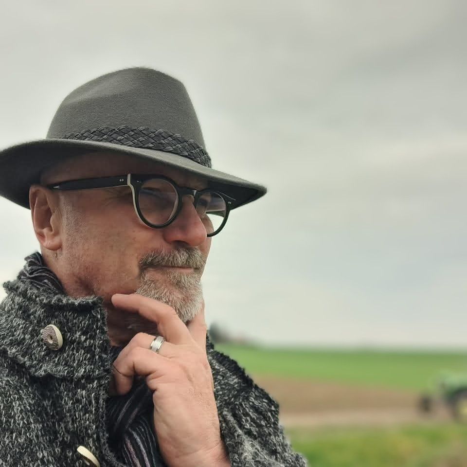 Ein Mann mit Hut und Brille steht nachdenklich auf einem Feld.
