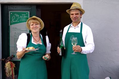 Zwei Personen in grünen Schürzen und Strohhüten halten Wein und Kuchen vor einem Gebäude.