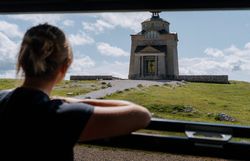 Eine Frau blickt von der Schneebergbahn auf das Elisabethkircherl am Hochschneeberg. Ein Kiesweg führt zur Kirche, die umgeben von einer grünen Wiese ist, was eine Atmosphäre der Erkundung und historischer Neugier erzeugt.