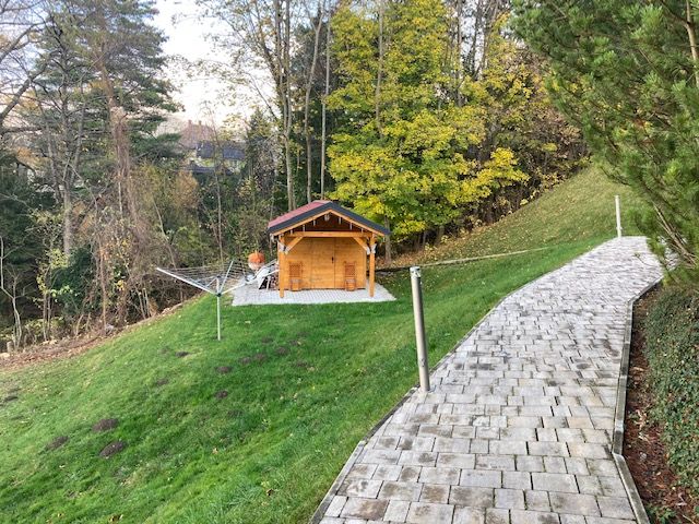 Ein Garten mit einem kleinen Holzhaus, umgeben von Bäumen und einem gepflasterten Weg.
