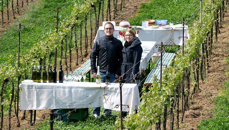 Zwei Personen stehen in einem Weinberg neben einem gedeckten Tisch mit Weinflaschen.