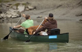 Zwei Personen und ein Hund in einem grünen Kanu auf einem Fluss.