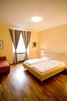 Ein gemütliches Doppelzimmer mit einem großen Bett, Holzboden, einem Sessel und einem Fenster mit Vorhängen.