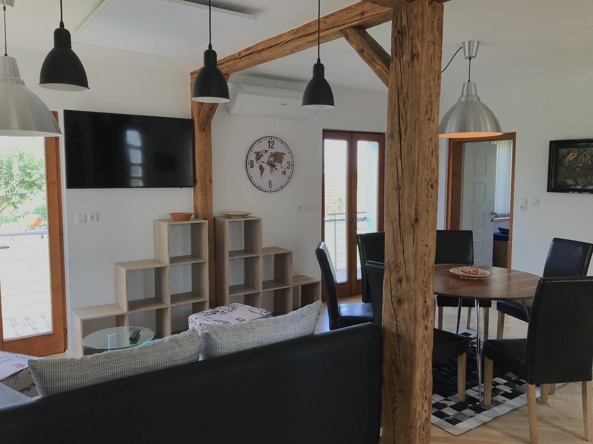 Moderner Wohnbereich mit Holzbalken, Esstisch, Stühlen, Wandregal, Uhr und Fernseher.