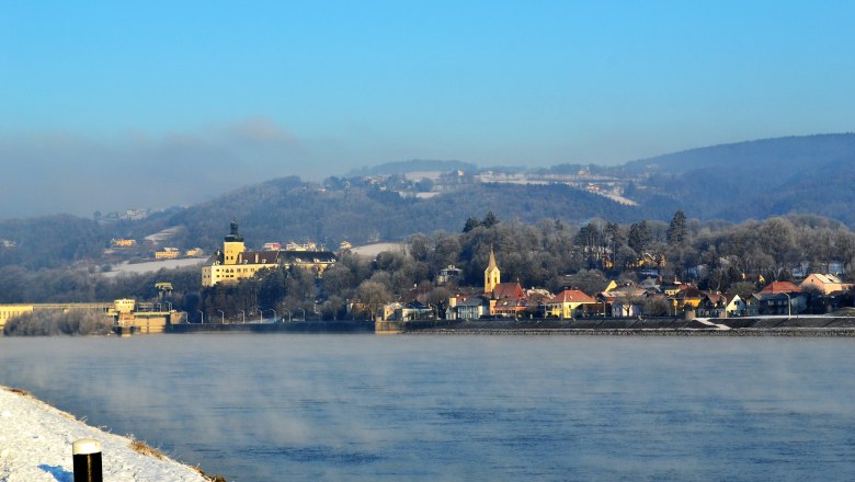 Winterlandschaft in Persenbeug mit Fluss, Gebäuden und Hügeln im Hintergrund.