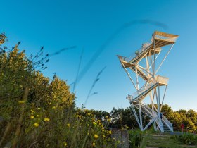 Der Aussichtsturm erhebt sich majest&auml;tisch &uuml;ber die umliegende Landschaft und bietet einen atemberaubenden Blick auf die sanften H&uuml;gel und dichten W&auml;lder. Die frische Bergluft und das Zwitschern der V&ouml;gel schaffen eine einladende Atmosph&auml;re f&uuml;r Wanderer und Naturliebhaber. Ein perfekter Ort, um die Sch&ouml;nheit der Natur in vollen Z&uuml;gen zu genie&szlig;en.