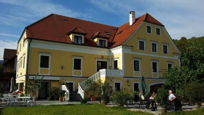 Gelbes Gebäude mit rotem Dach und Terrasse, umgeben von Bäumen.