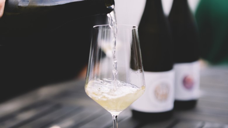 Wei&szlig;wein wird in ein Glas auf einem Holztisch eingeschenkt, mit zwei Weinflaschen im Hintergrund.