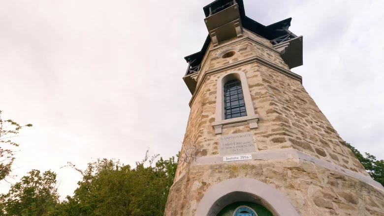 Steinerner Aussichtsturm mit Fenstern und Inschrift, umgeben von Bäumen.