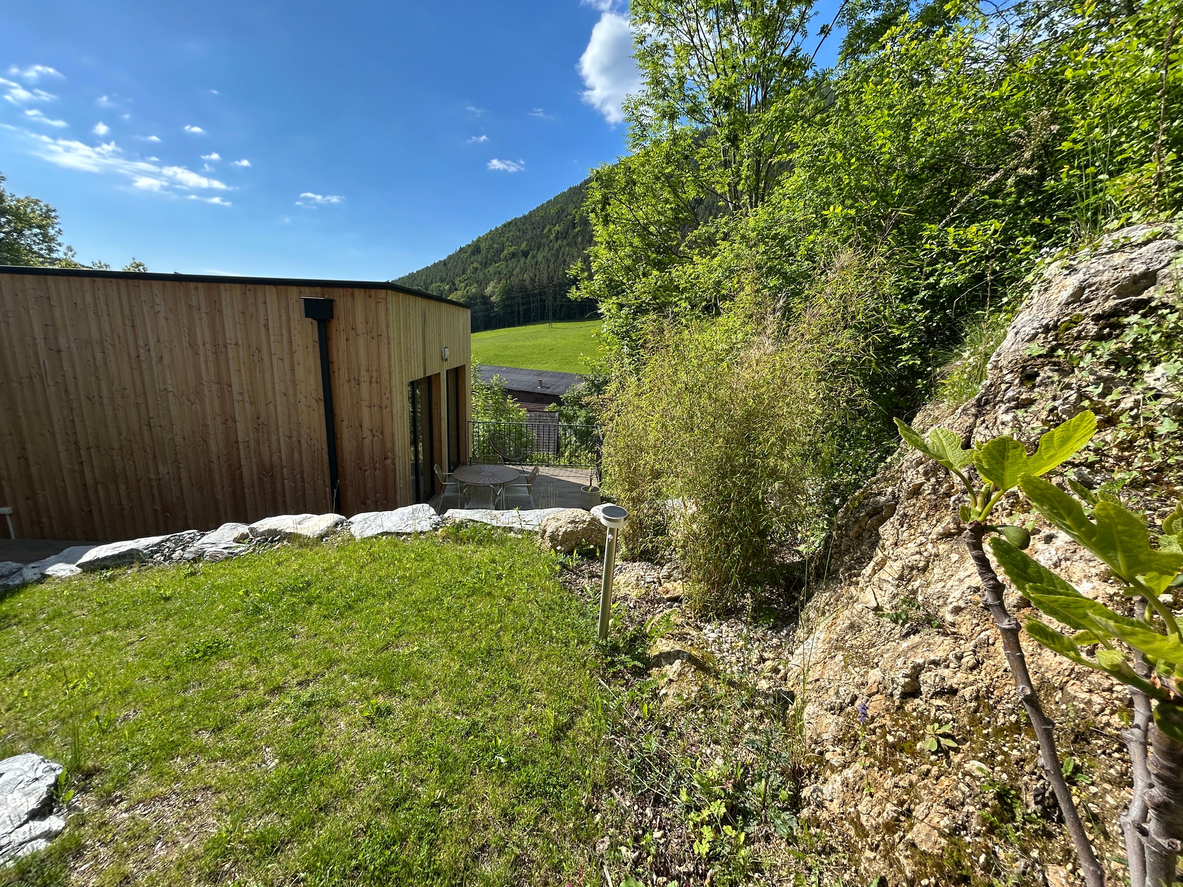 Ein modernes Tiny House mit Holzverkleidung steht in einer grünen, hügeligen Landschaft. Im Vordergrund sind Felsen und Pflanzen zu sehen.