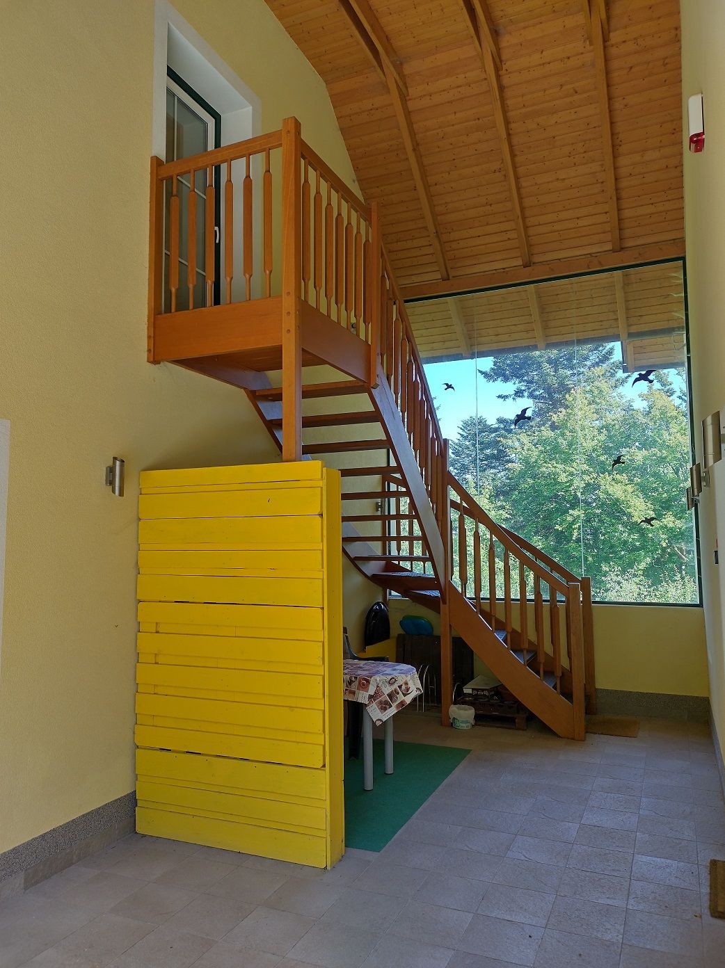 Holztreppe in einem Innenraum mit gelber Wand und großem Fenster mit Blick auf Bäume.