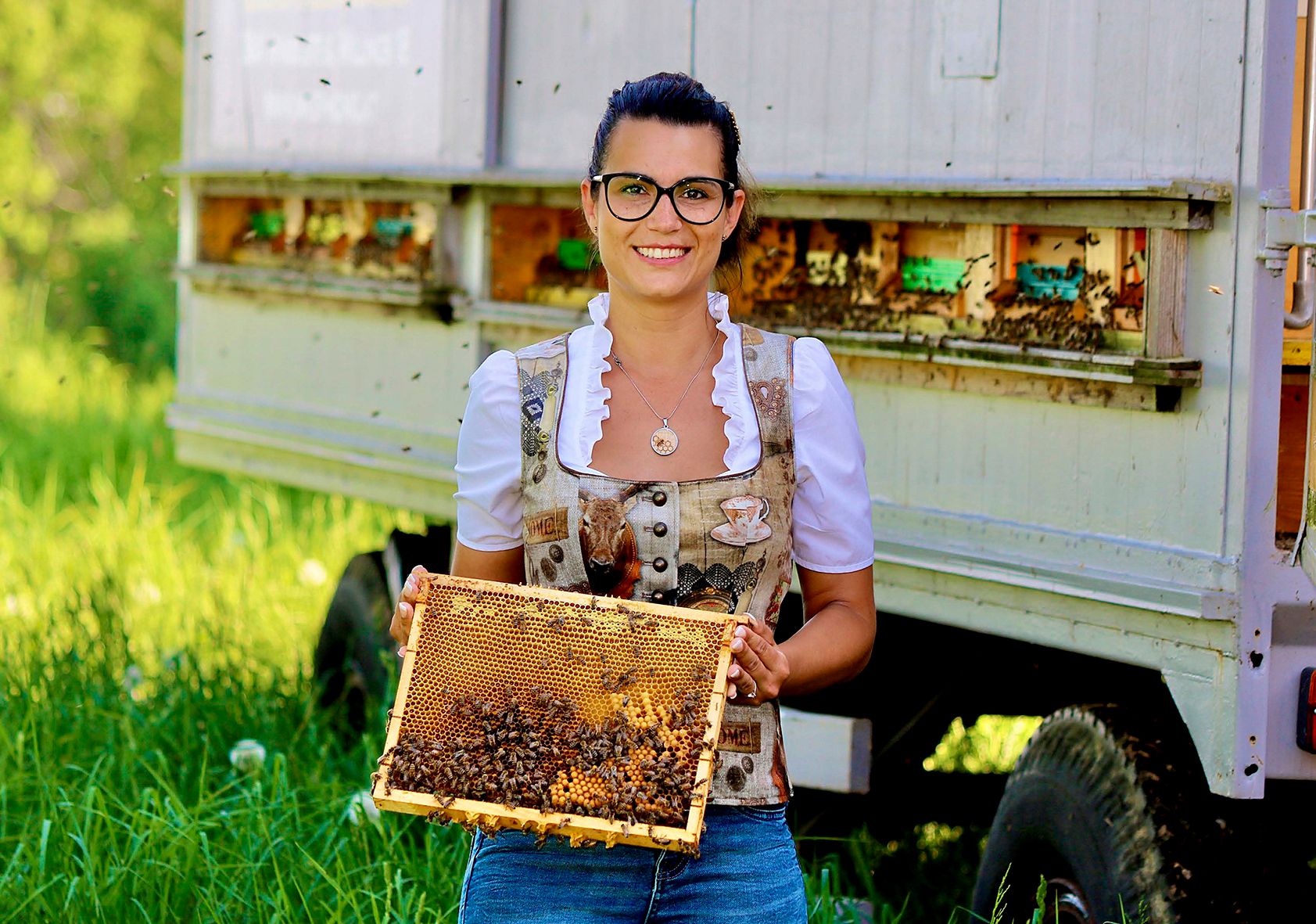 Eine Frau in traditioneller Kleidung hält eine Bienenwabe vor einem Bienenwagen.