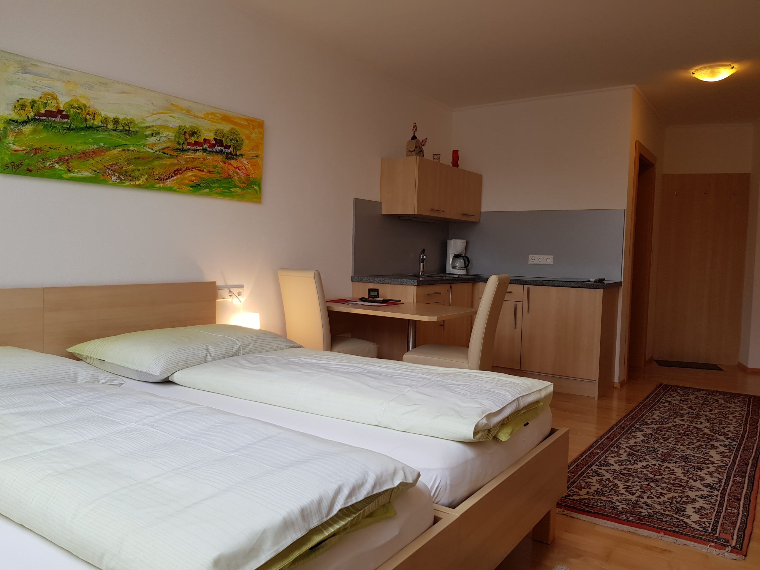 Ein modernes Zimmer mit zwei Betten, einem Schreibtisch, einer kleinen Küchenzeile und einem bunten Gemälde an der Wand.