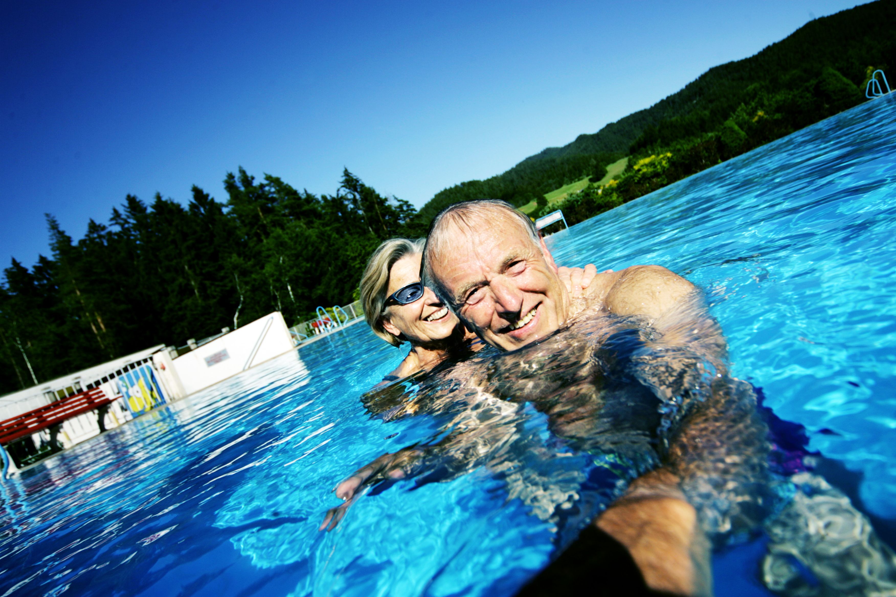 Ein älteres Paar schwimmt lächelnd in einem Freibad mit Wald im Hintergrund.