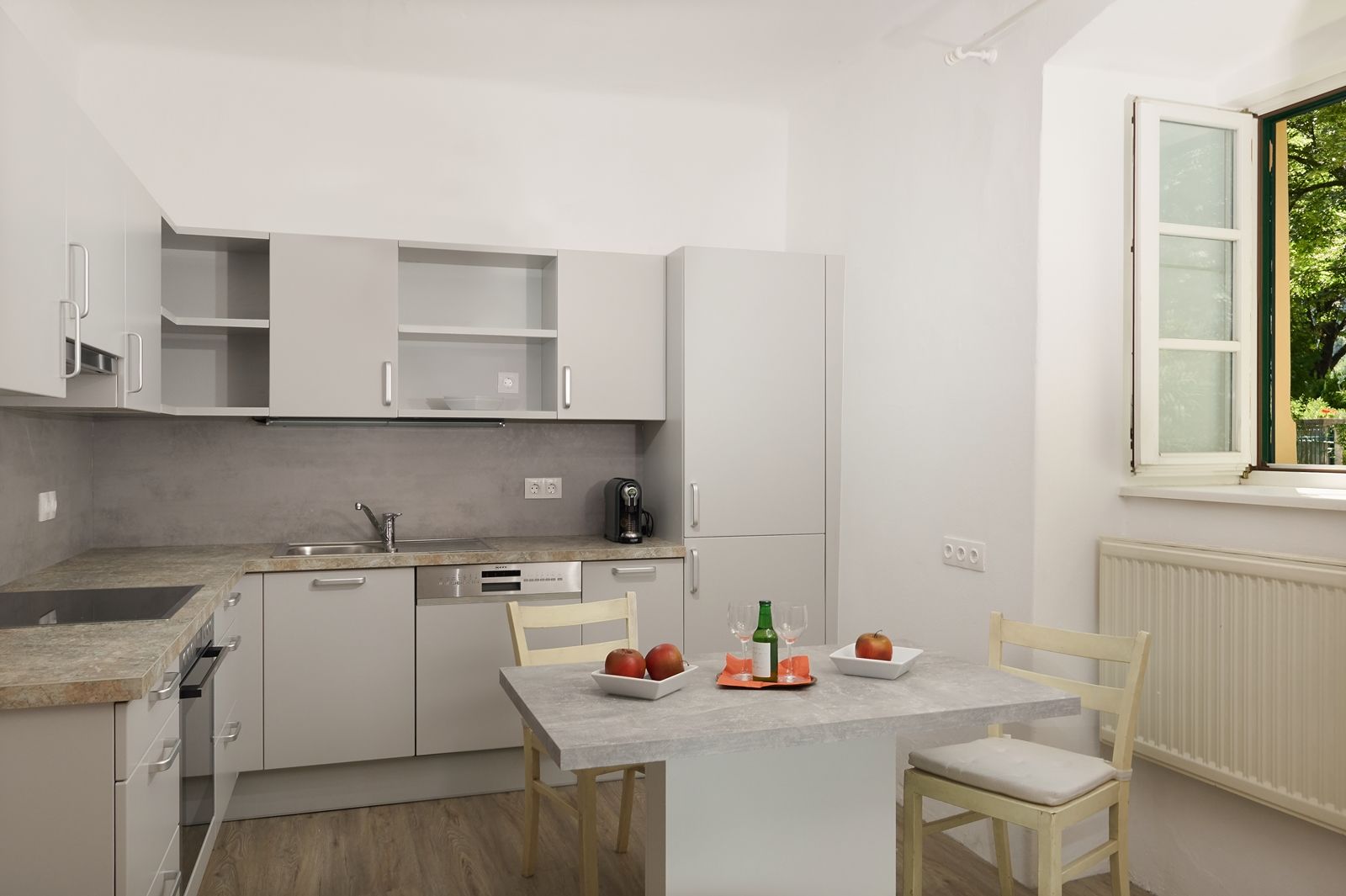 Moderne Küche mit grauen Schränken, Tisch mit Äpfeln und Getränk, offenes Fenster.