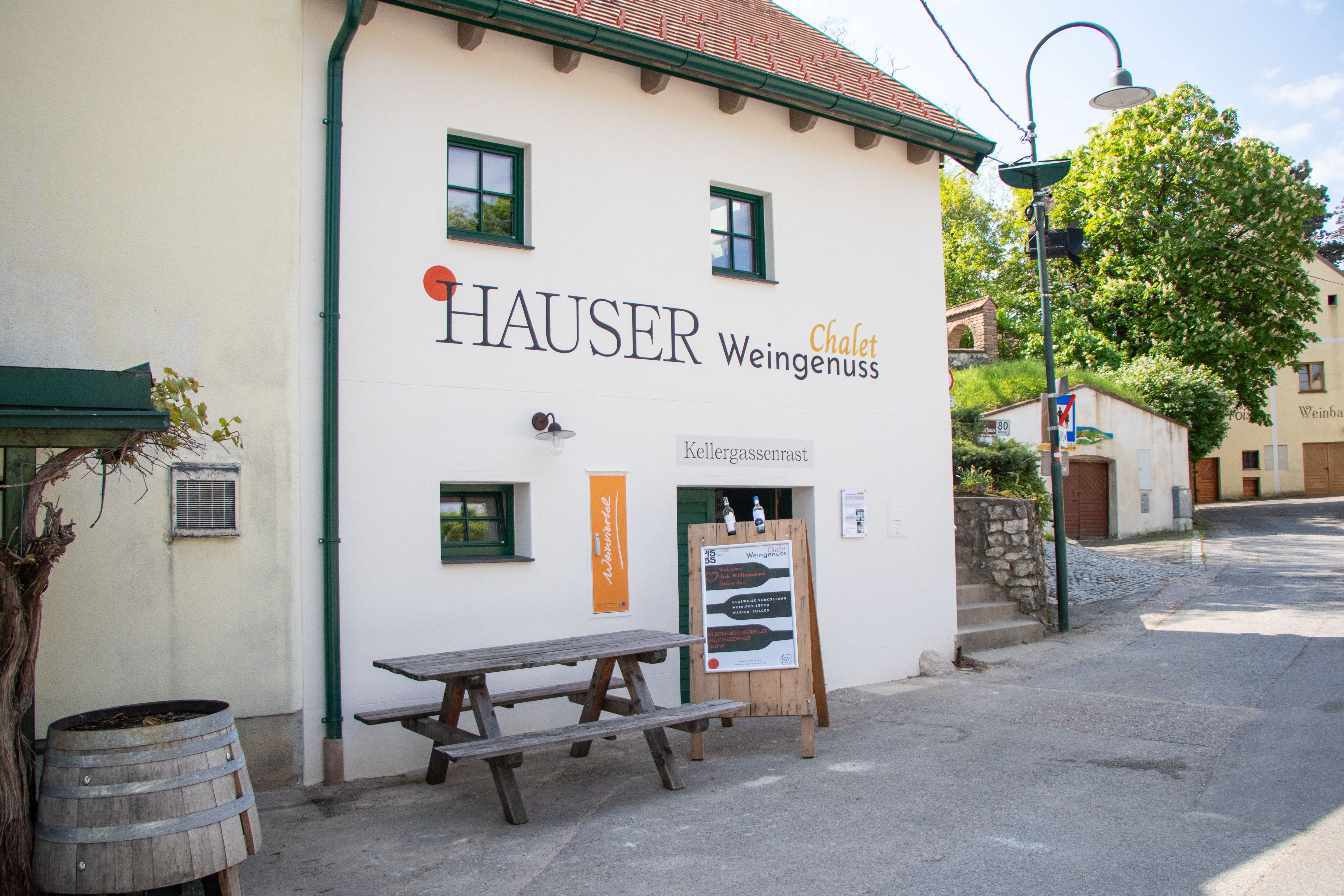 Außenansicht eines Gebäudes mit der Aufschrift 'Hauser Chalet Weingenuss'. Vor dem Gebäude steht ein Holztisch und ein Weinfass.