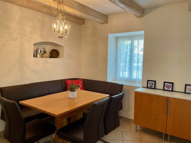 Ein gemütlicher Essbereich mit Holztisch, schwarzen Stühlen und Eckbank. An der Wand hängt ein Kronleuchter, daneben ein Fenster und ein Sideboard.