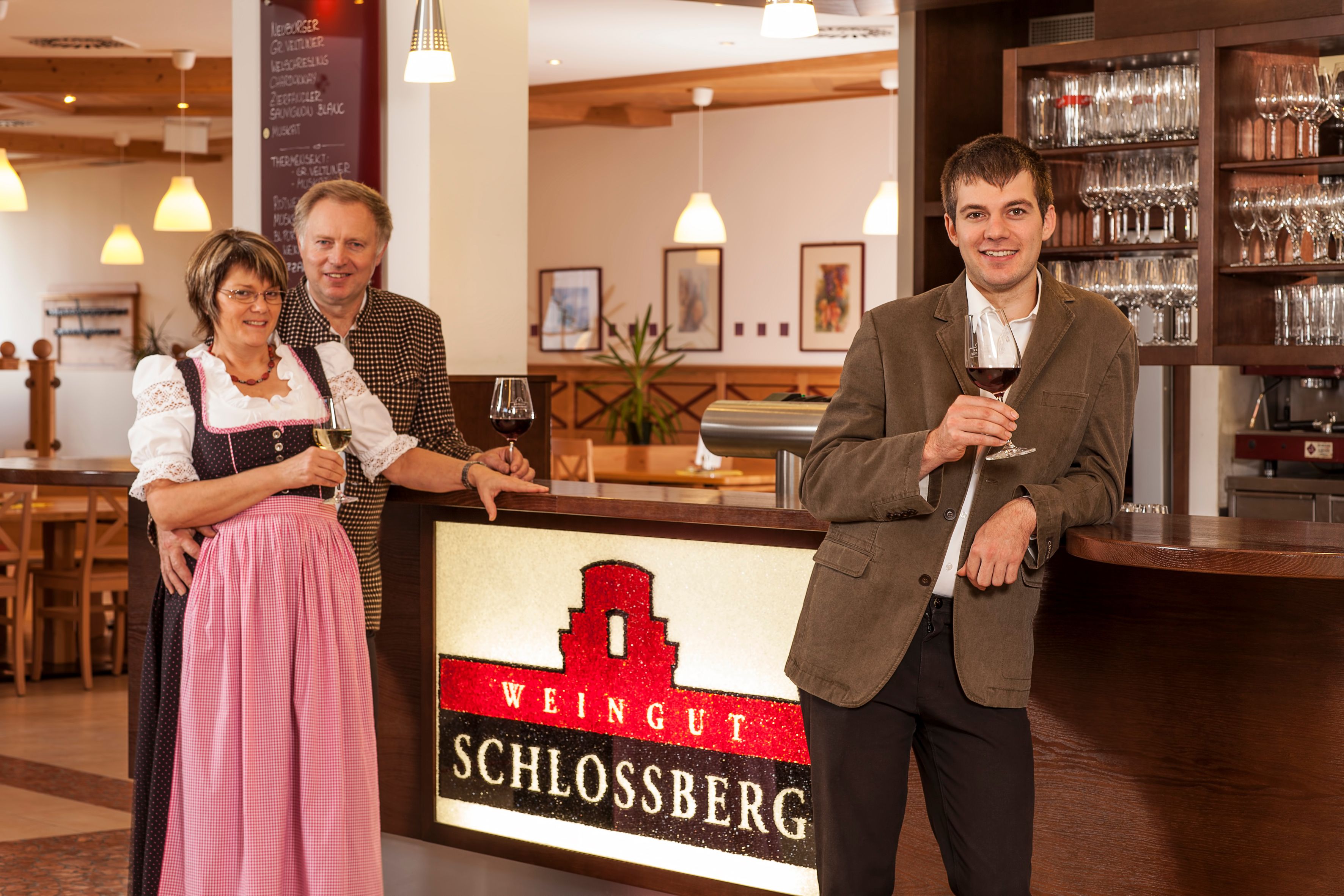 Drei Personen stehen an einer Bar im Weingut Schlossberg mit Weingläsern in der Hand.