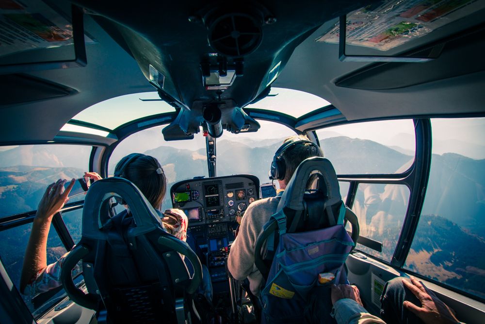 Blick aus dem Cockpit eines Helikopters mit zwei Personen, die über eine Berglandschaft fliegen.