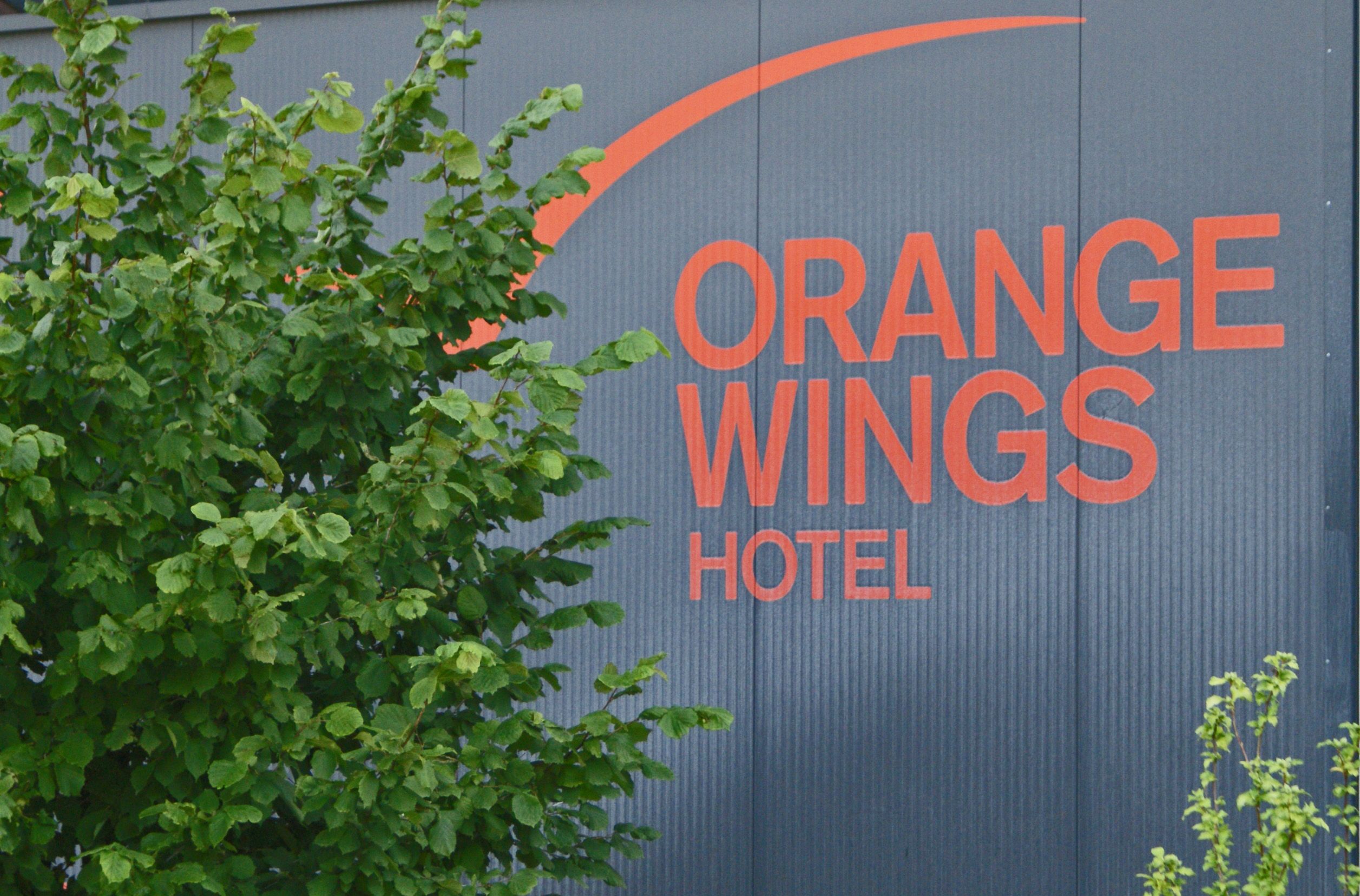 Logo des Orange Wings Hotels auf einer Wand mit Pflanzen im Vordergrund.