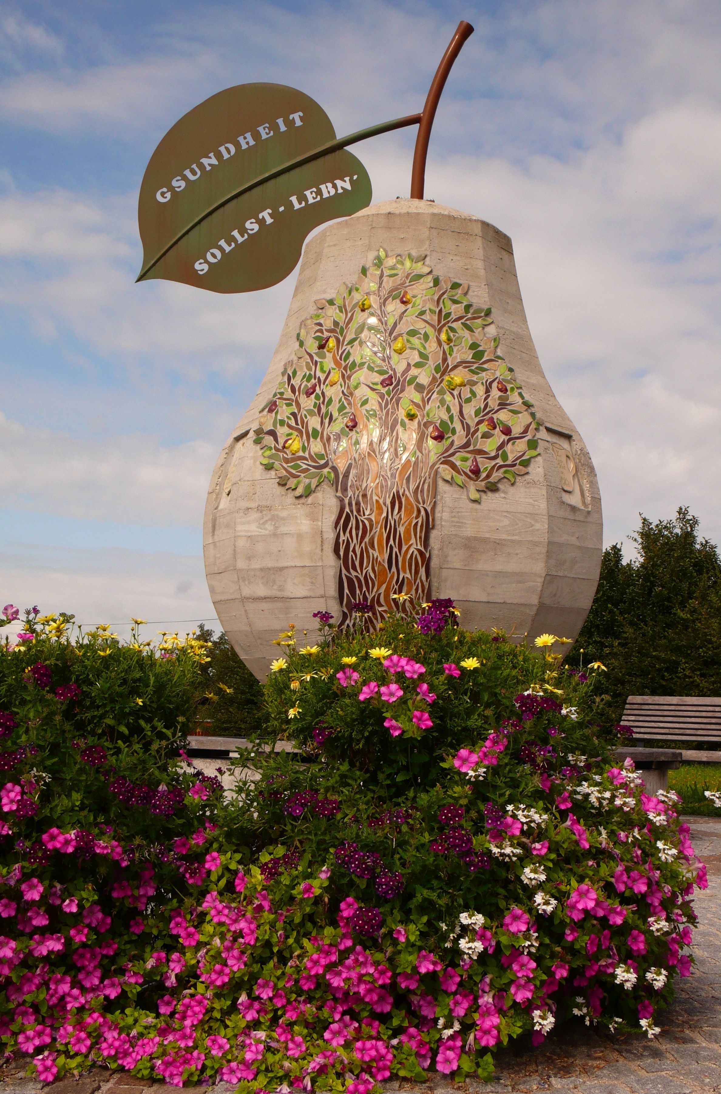 Große Betonbirne mit Blattaufschrift 'Gsundheit sollst lebn' und Blumen im Vordergrund.