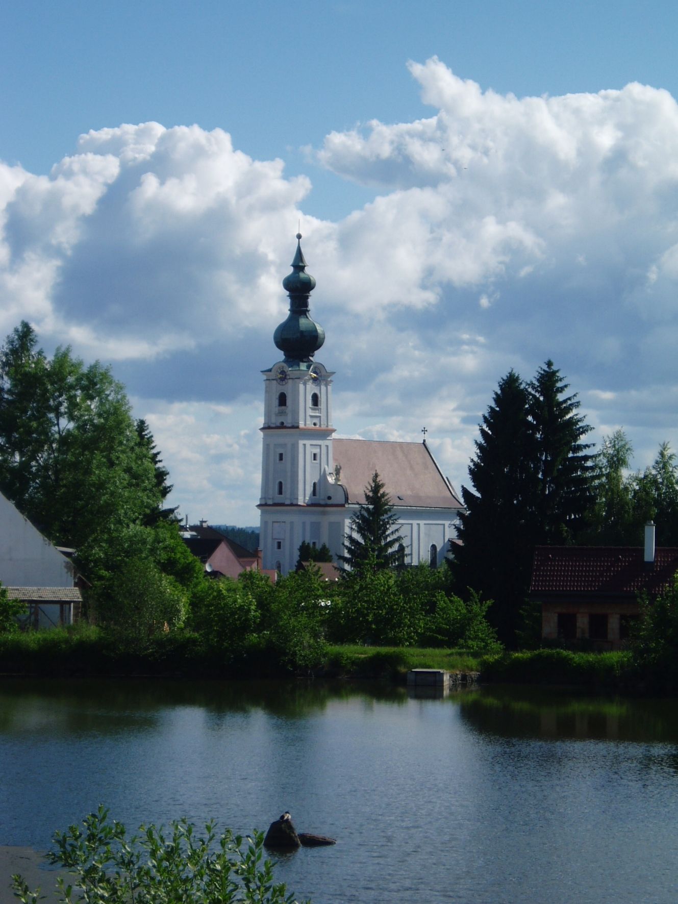 Kirche in Kirchberg am Walde mit Teich im Vordergrund und bewölktem Himmel.