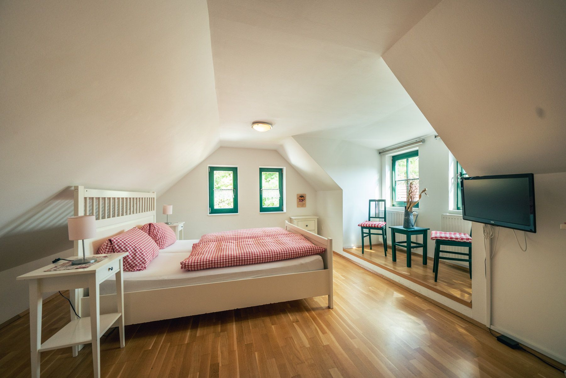 Helles Schlafzimmer mit Dachschräge, Doppelbett, Holzboden, zwei Fenstern, Tisch mit Stühlen und Fernseher an der Wand.
