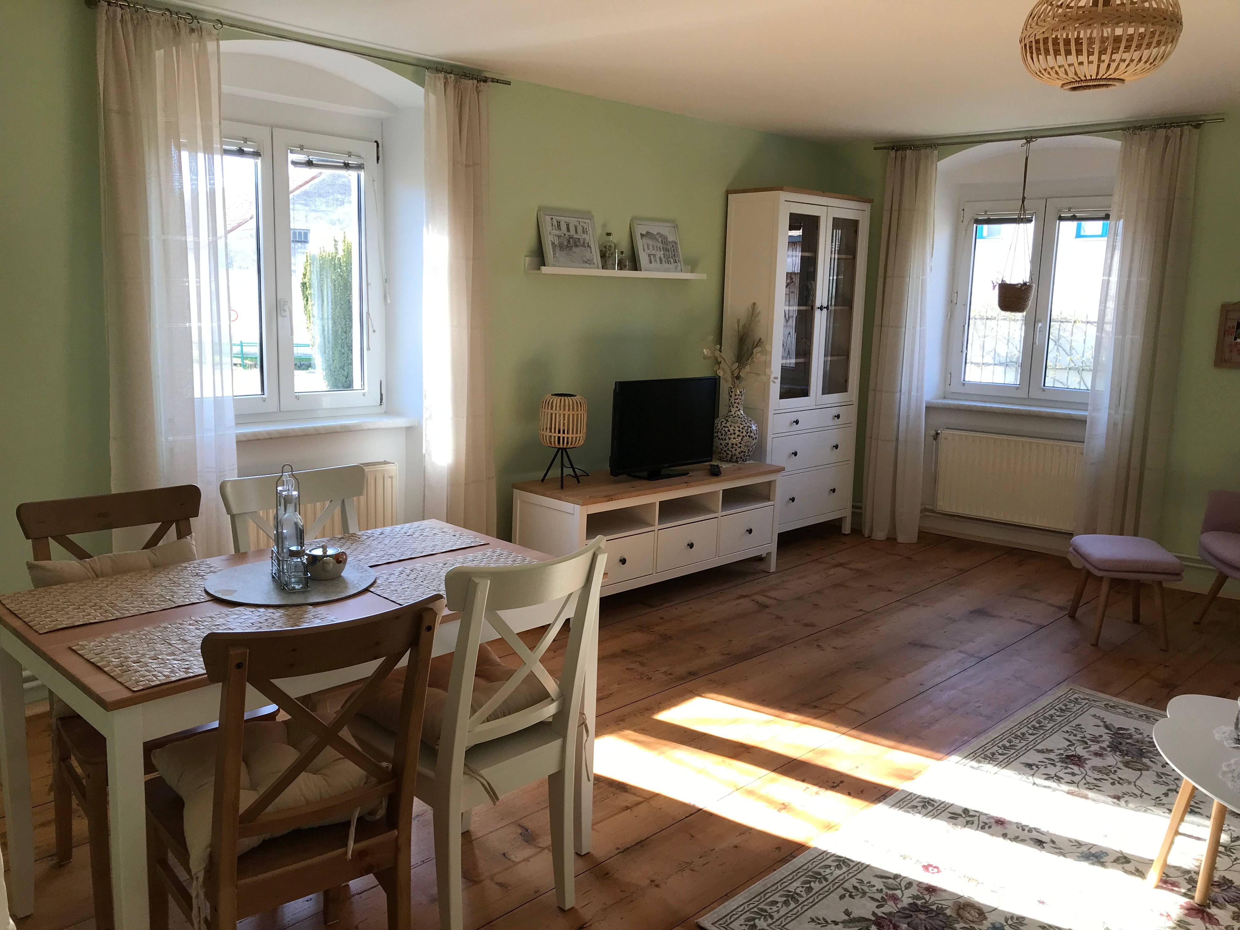 Heller Raum mit Esstisch, Stühlen, TV-Schrank und großen Fenstern.