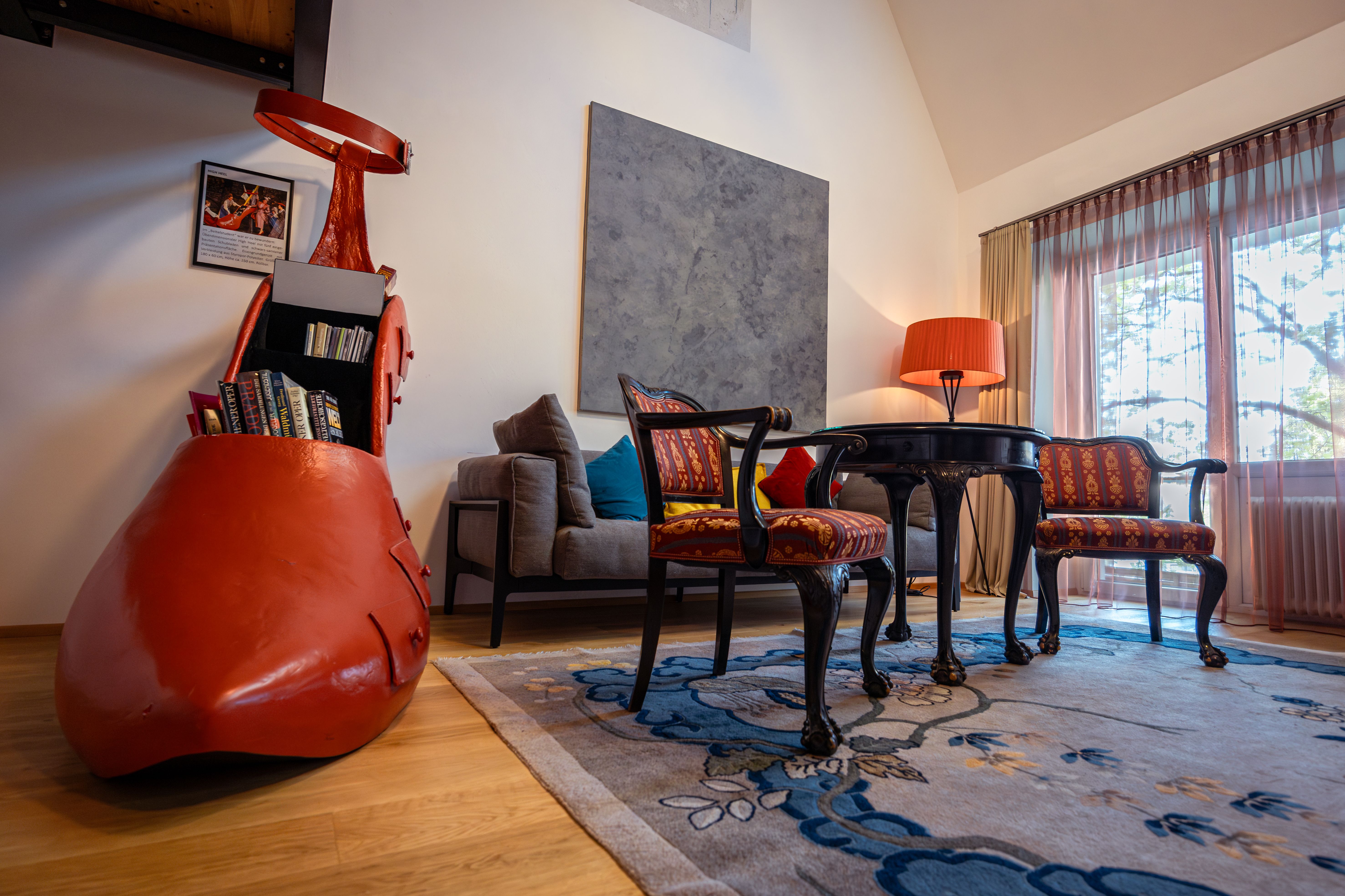 Ein stilvoll eingerichtetes Hotelzimmer mit einem großen roten Schuh als Bücherregal, antiken Stühlen, einem Tisch und einer Stehlampe.