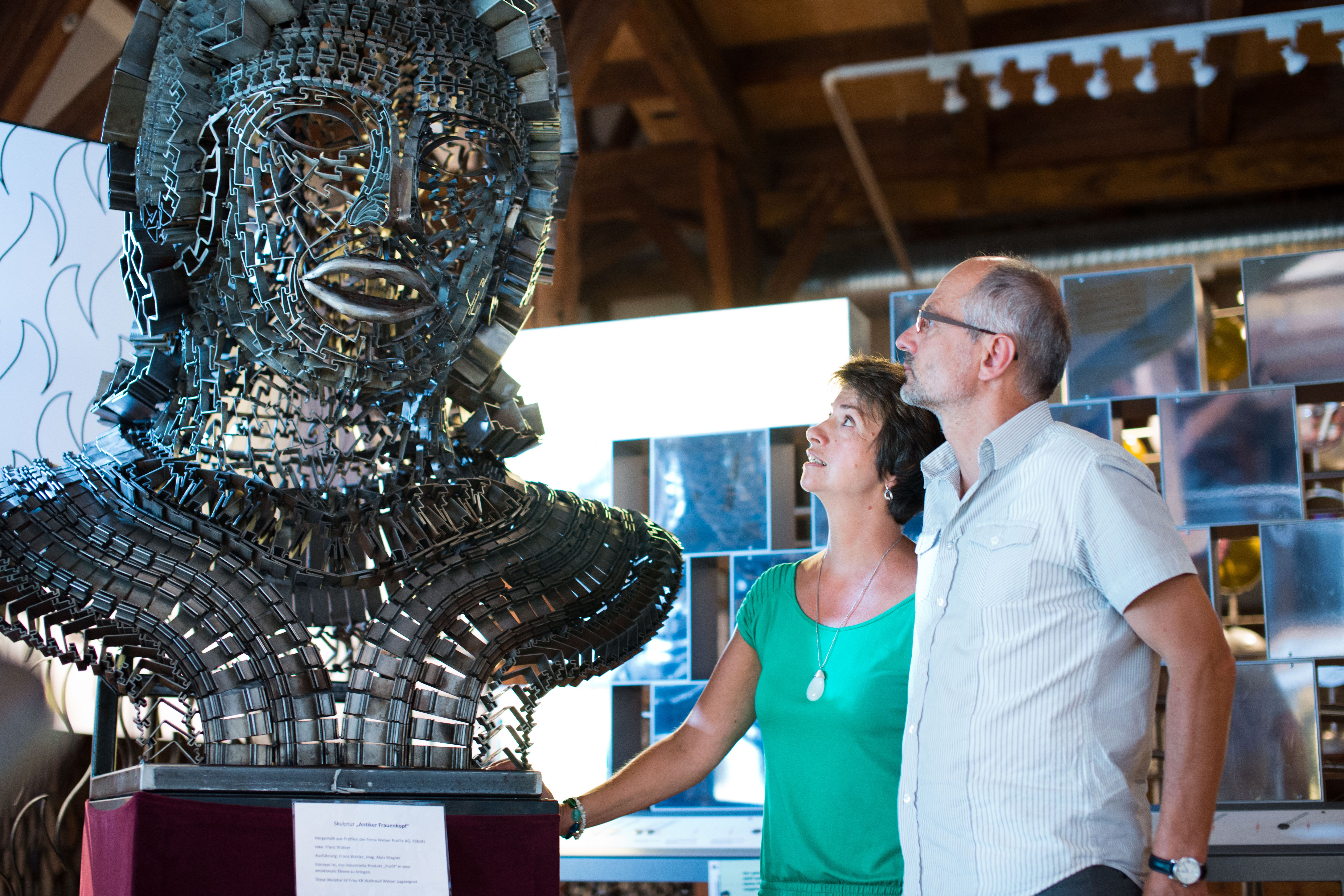Zwei Personen betrachten eine große Metallskulptur in einem Ausstellungsraum.