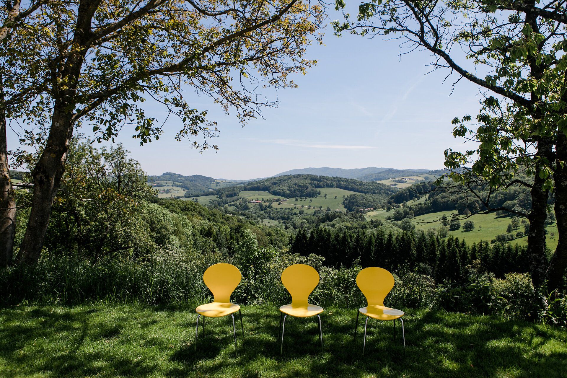 Drei leuchtend gelbe Stühle laden dazu ein, die atemberaubende Aussicht auf die sanften Hügel und das üppige Grün der Umgebung zu genießen. Die ruhige Atmosphäre und die frische Bergluft schaffen einen perfekten Ort für eine entspannte Auszeit in der Natur. Hier, umgeben von Bäumen und der malerischen Landschaft, wird der Sommer in den Bergen zum unvergesslichen Erlebnis.