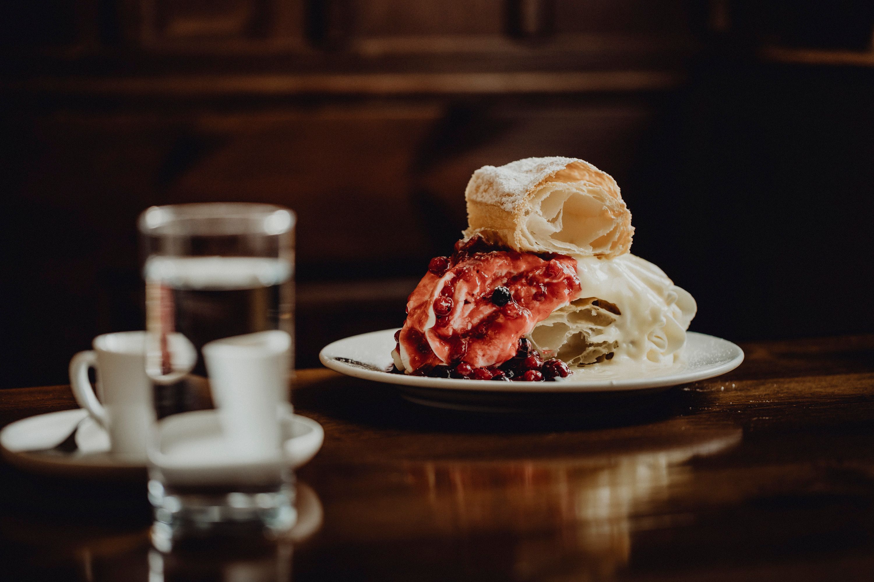 Ein Teller mit einer Klostercremeschnitte, garniert mit roten Beeren, steht auf einem Holztisch. Im Hintergrund sind ein Glas Wasser und zwei Espressotassen zu sehen.