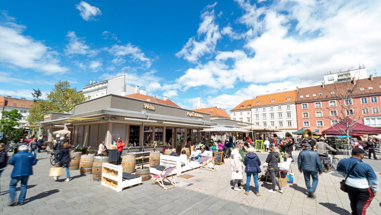 Ein belebter Marktplatz mit Menschen, die an Ständen und Cafés verweilen, unter einem blauen Himmel.