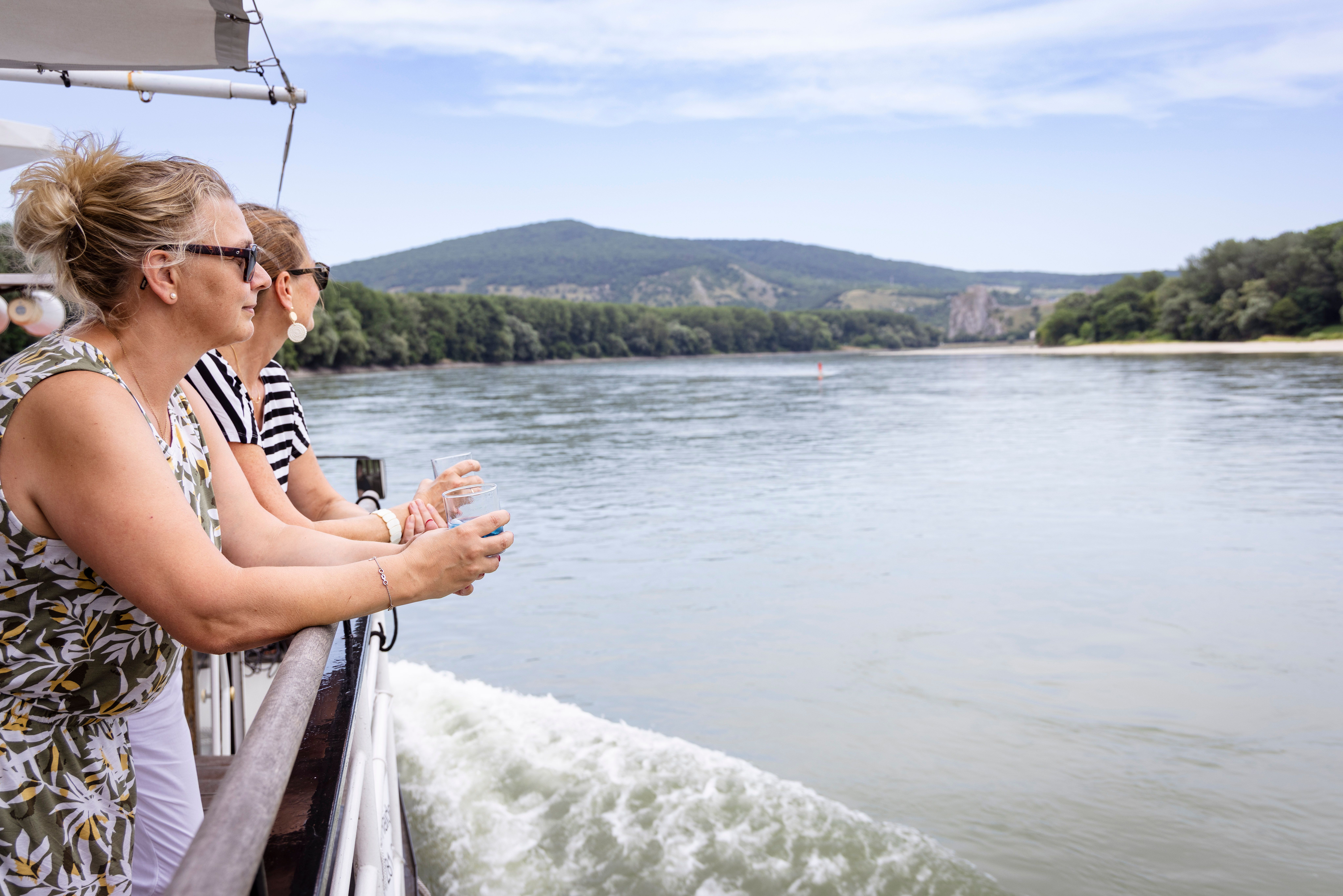 Zwei Frauen stehen an der Reling eines Schiffes auf der Donau und genießen die Aussicht auf die umliegende Landschaft.