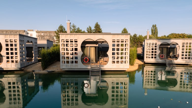 Therme Laa | Hotel | Silent Spa | Villas, © Niederösterreich Werbung / Maximilian Pawlikowsky Moderne Villen auf Wasser mit reflektierenden Fassaden und Treppen, umgeben von Natur.