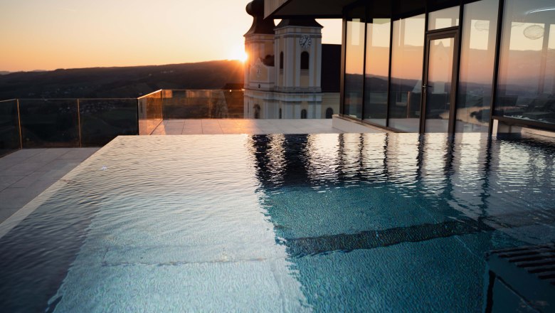 Infinitypool bei Sonnenuntergang mit Blick auf ein Geb&auml;ude und die Landschaft.