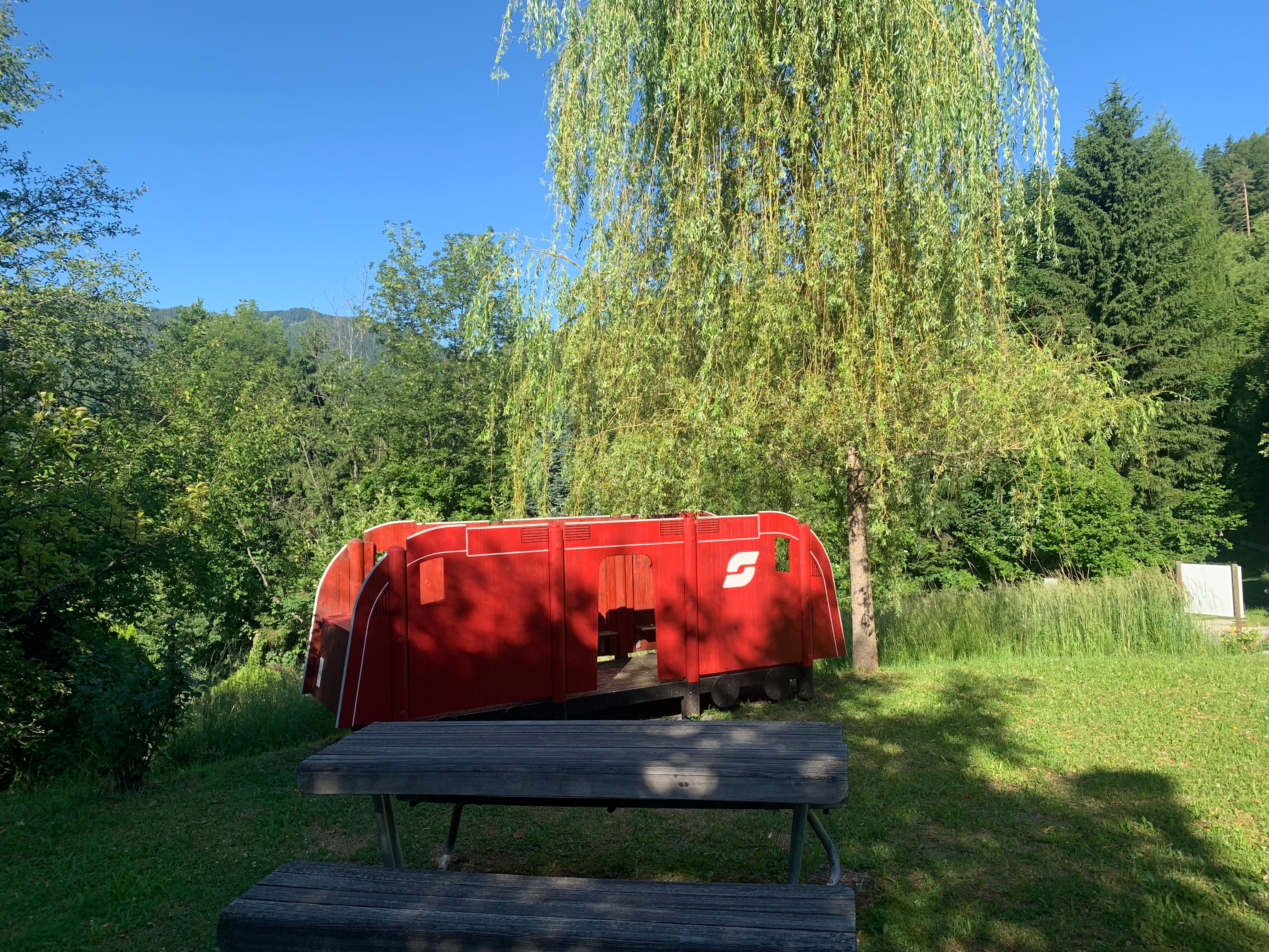 Rote Spielzeuglokomotive im Freien neben einem Baum und einer Holzbank.