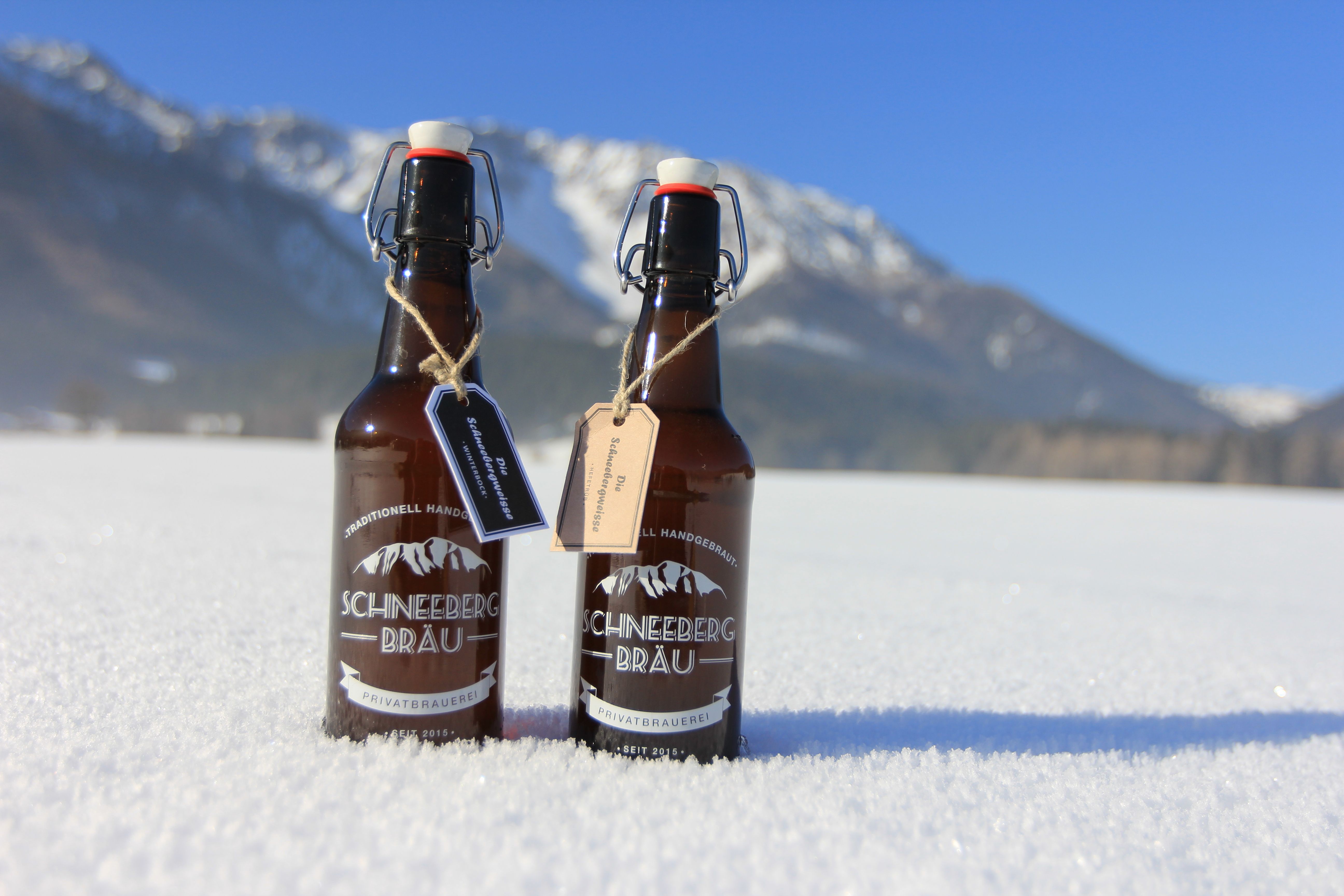Zwei Bierflaschen der Marke Schneeberg Bräu stehen im Schnee vor einer Bergkulisse.
