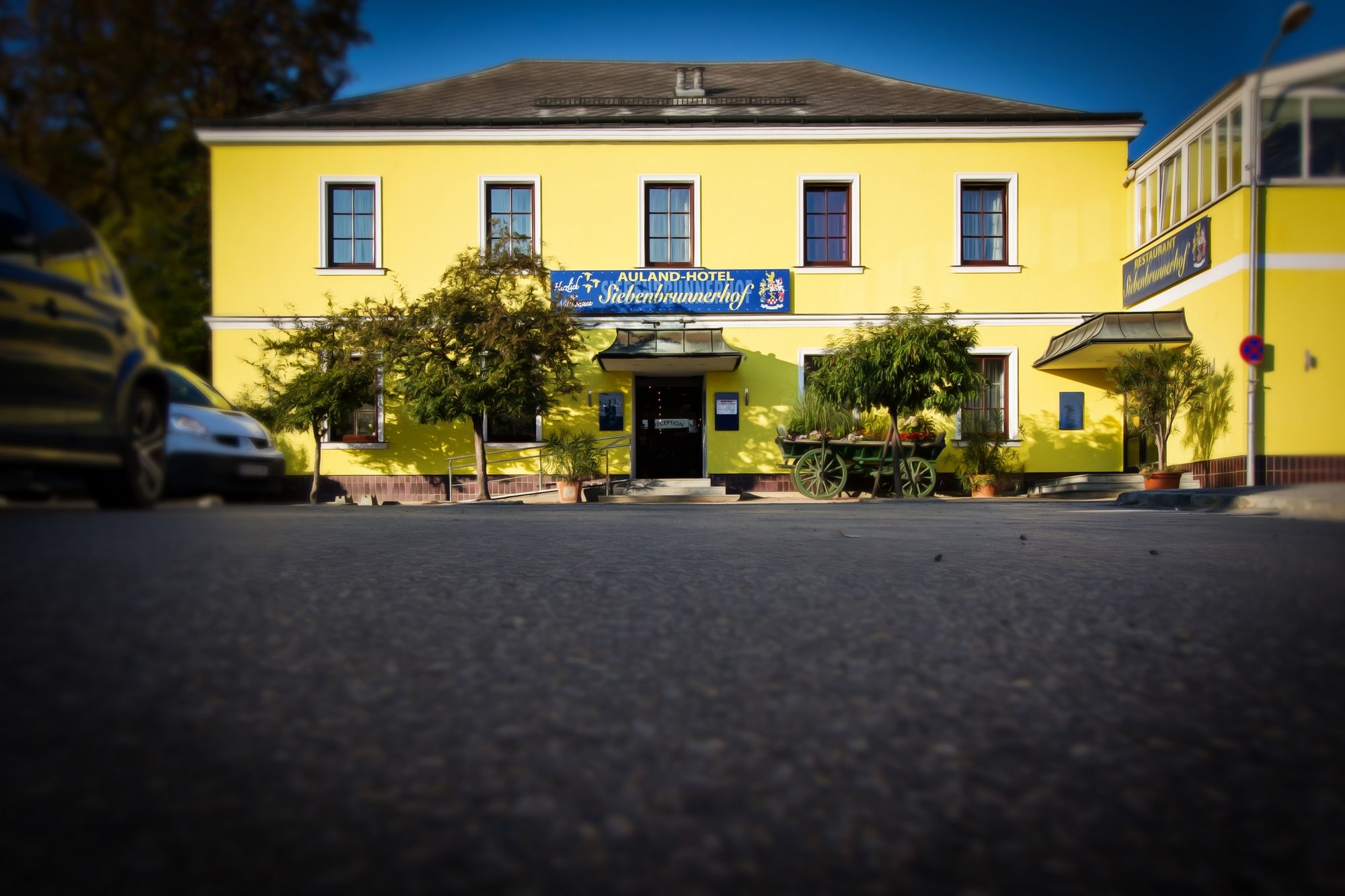 Gelbes Gebäude mit der Aufschrift 'Auland-Hotel Siebenbrunnerhof', umgeben von Bäumen und einem alten Wagen.