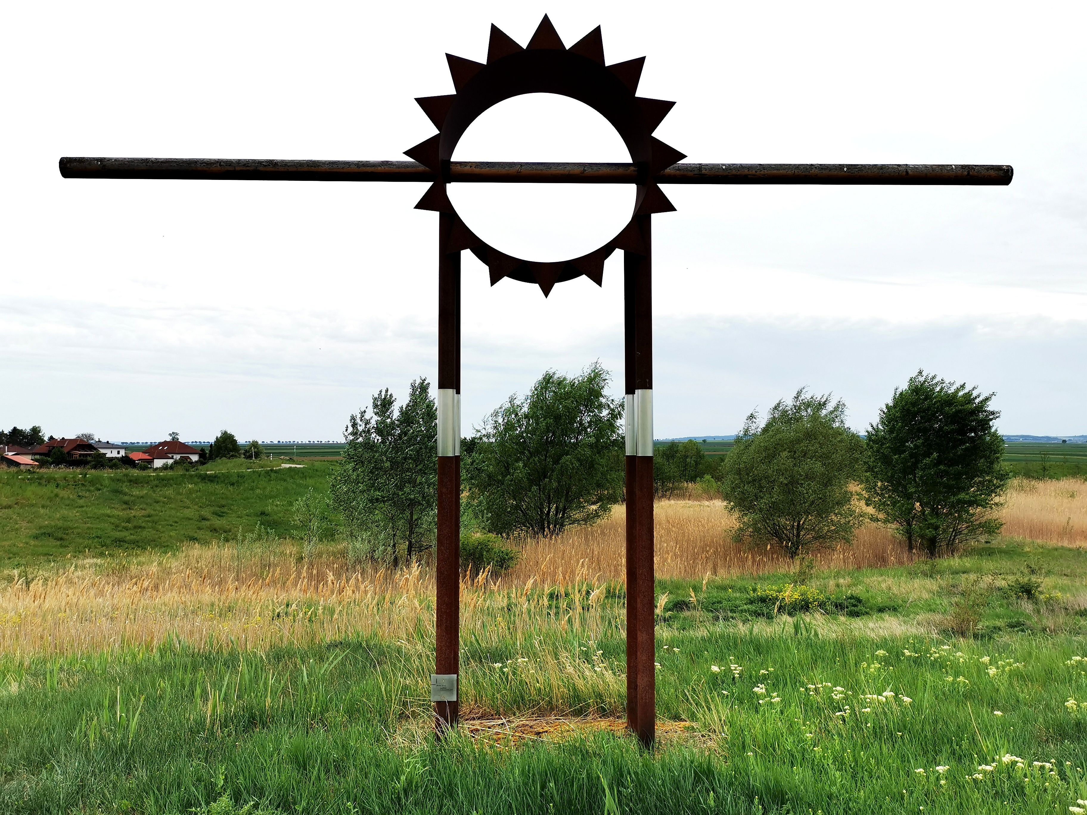 Metallskulptur in Form einer Sonne auf einer Wiese mit Bäumen im Hintergrund.