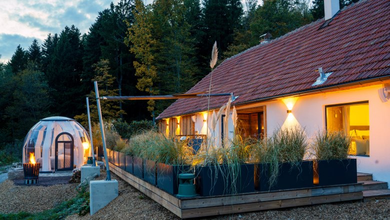 Chalet´s & Plätze voller WUNDER - Weidehaus De Luxe, © WUNDERVOLL eben Ein modernes Chalet mit Terrasse und einem transparenten Kuppelzelt im Wald bei Dämmerung.