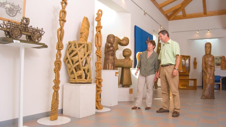 Zwei Personen betrachten Holzskulpturen in einem Ausstellungsraum mit Holzbalkendecke.