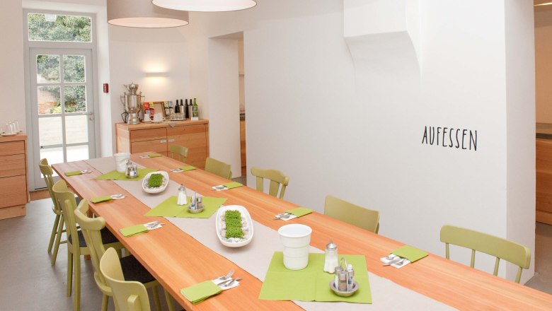 Ein heller Speiseraum mit einem langen Holztisch, gr&uuml;nen St&uuml;hlen und Dekoration. An der Wand steht 'AUFESSEN'.