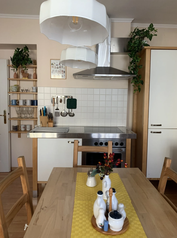 Gemütliche Küche mit Holzmöbeln, Esstisch und Stühlen, zwei Fenstern mit Vorhängen, und Küchenutensilien auf der Arbeitsfläche.