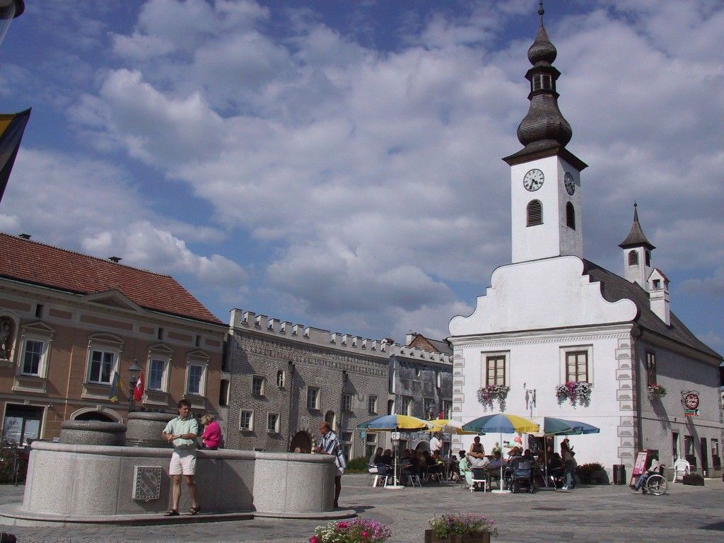 Ein Stadtplatz mit einem weißen Gebäude mit Turm und Uhr, Menschen sitzen unter Sonnenschirmen, ein Brunnen im Vordergrund.