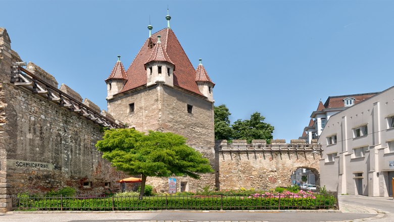 Historischer Reckturm mit rotem Dach und angrenzender Stadtmauer in einer urbanen Umgebung.